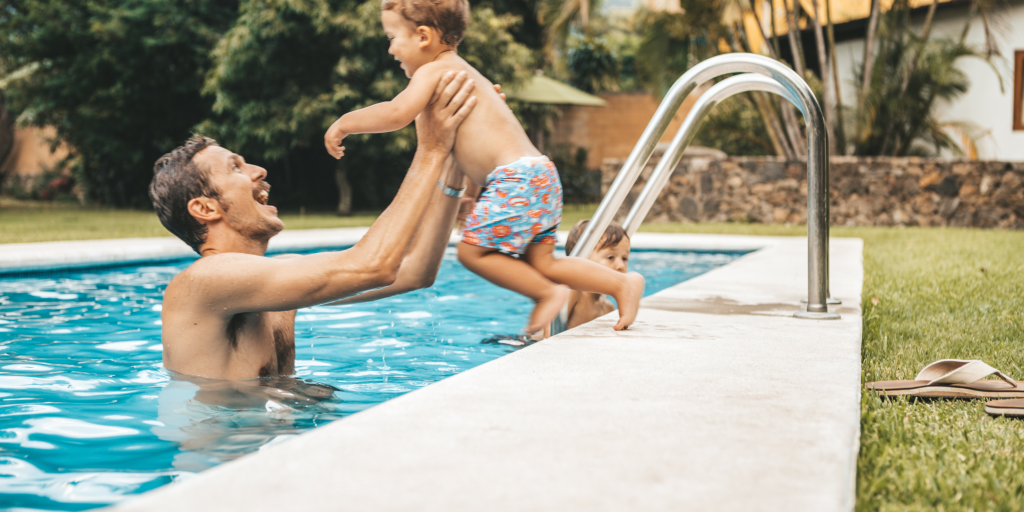 Adult lifting a toddler out of a backyard swimming pool while another child watches nearby.