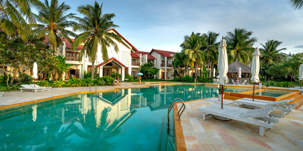 Resort-style swimming pool surrounded by palm trees, lounge chairs, and tropical villas on a clear sunny day.