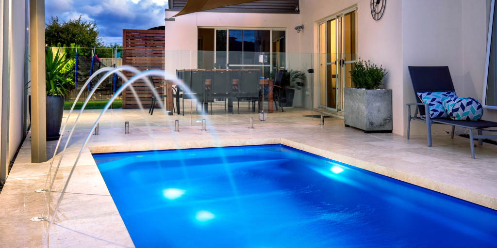 Modern backyard swimming pool at dusk with illuminated water, sleek deck, water features, and outdoor seating.