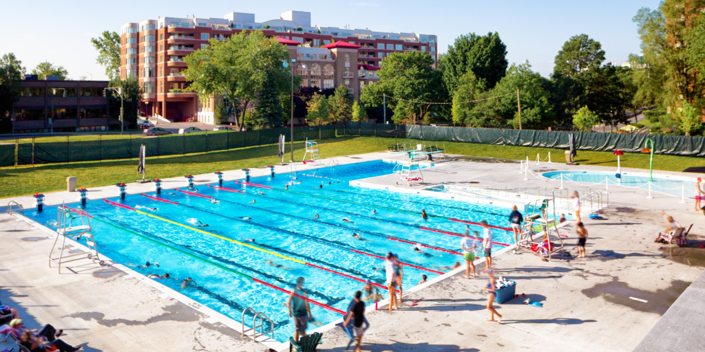Large outdoor lap pool with multiple swim lanes, swimmers in the water, and people gathered on the pool deck in a city park setting.