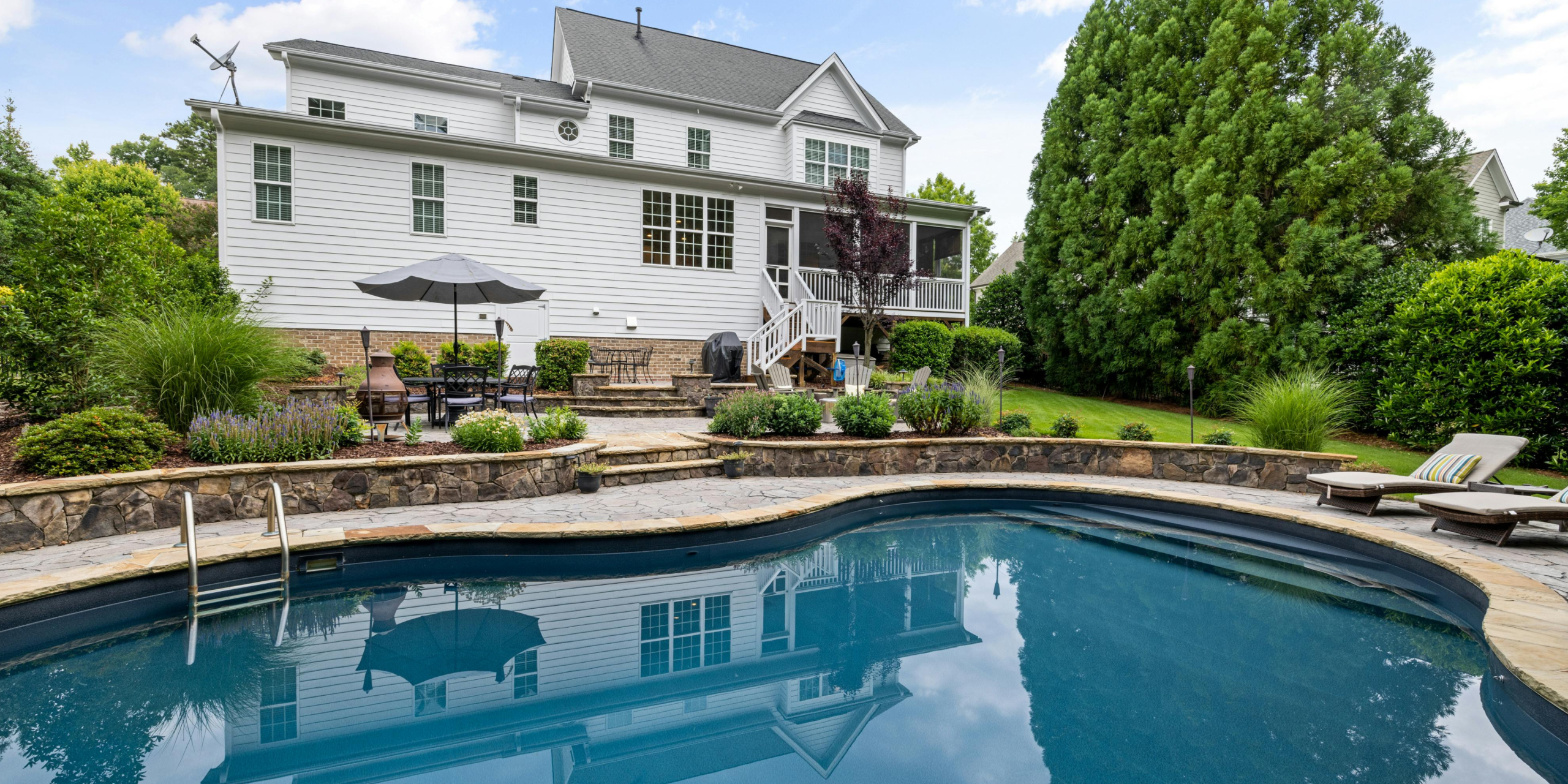 Backyard residential pool in front of a large white home surrounded by landscaped gardens.