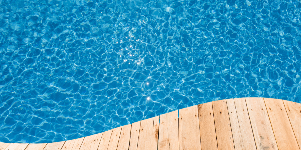 Clear blue swimming pool water with sunlight reflections on the surface near a wooden pool deck.
