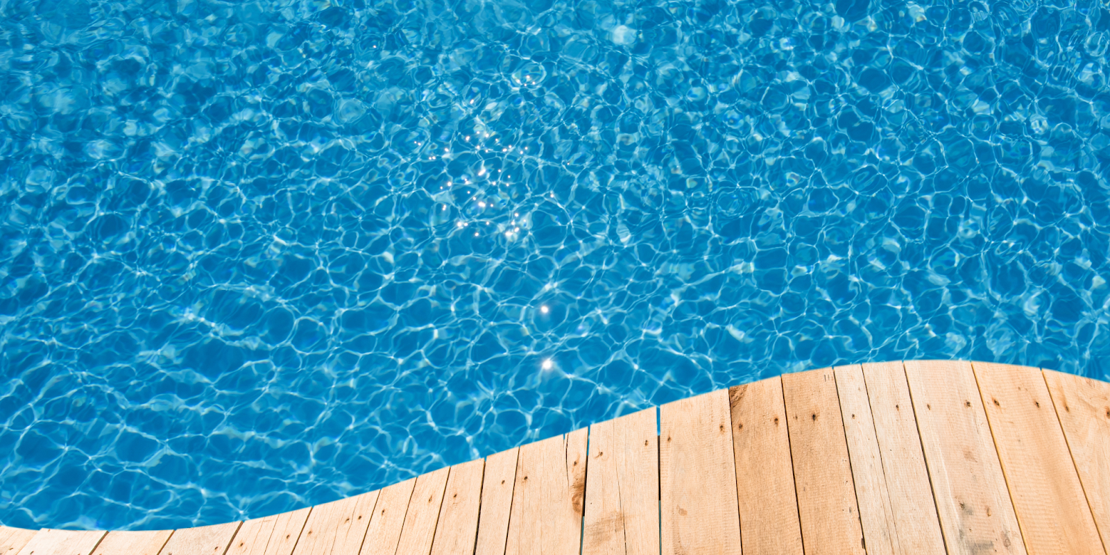 Clear blue swimming pool water with sunlight reflections on the surface near a wooden pool deck.