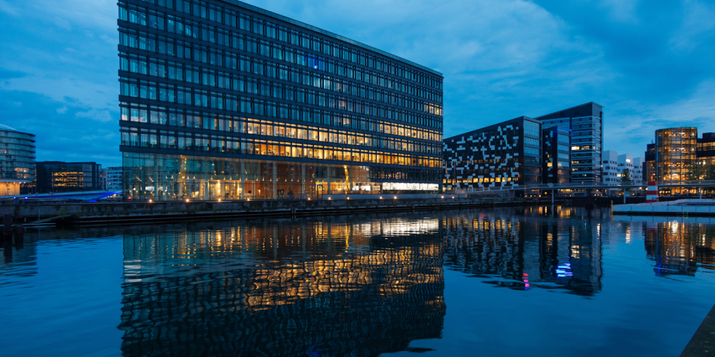 Modern glass office building on the waterfront at dusk with lights reflecting on calm water.