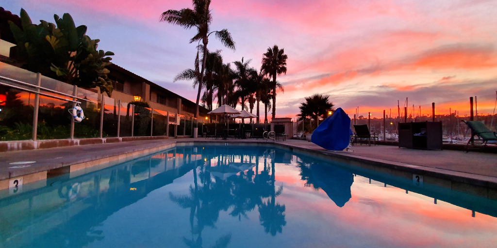 Outdoor swimming pool at sunset with palm trees, lounge chairs, and boats docked in a marina in the background.