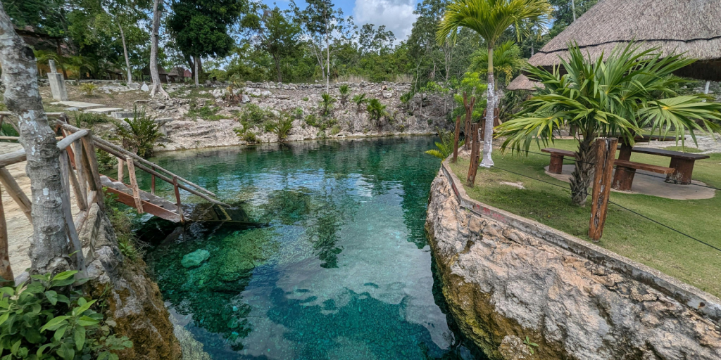 Natural outdoor pool with clear turquoise water surrounded by stone walls, tropical plants, and wooden railings.